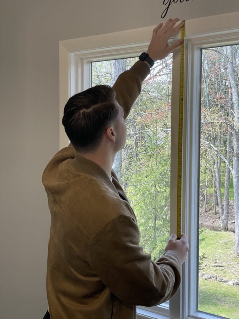 A man measures a window with a tape measure, facing a wooded area outside, as he prepares to install custom curtains.